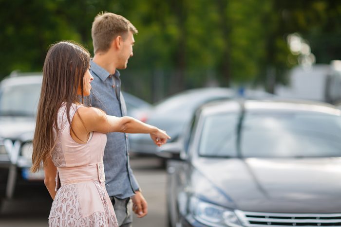 Beautiful young couple standing at the dealership choosing the car to buy. A beautiful sunny day to buy a new car. Happy beautiful couple is choosing a new car at dealership. Parking for sale.