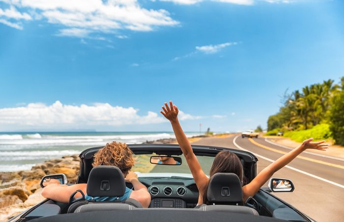 Road trip car holiday happy couple driving convertible car on summer travel Hawaii vacation. Woman with arms up having fun, young man driver