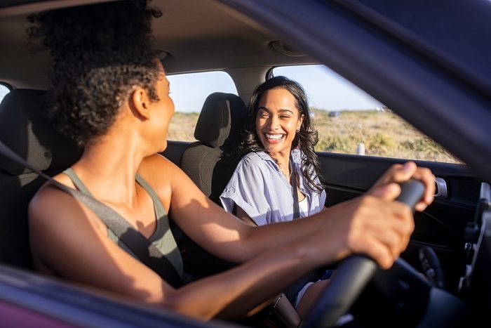 Beautiful latin hispanic woman with friend enjoying and laughing in car while going on a road trip. Cheerful girl friends enjoying trip and drive in car at sunset. Black woman driving a car.